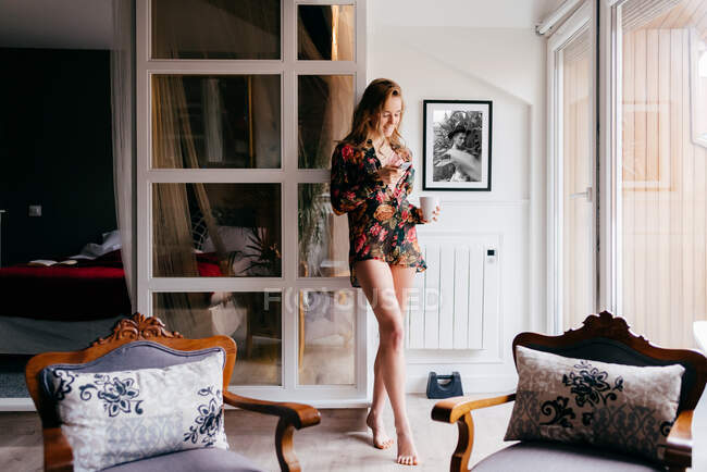 Bastante delgada joven hembra en colorida túnica de seda bebiendo café mientras está de pie frente a la ventana en la acogedora sala de estar por la mañana utilizando el teléfono móvil - foto de stock