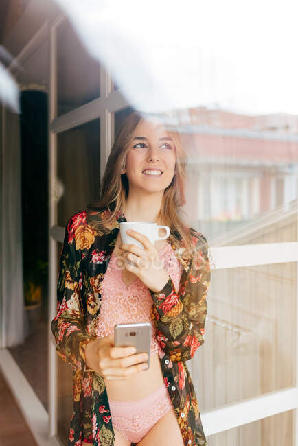 Feliz atractiva joven hembra en lencería elegante y colorida bata mirando a la ventana mientras pasa la mañana con una taza de café y teléfono inteligente en casa - foto de stock