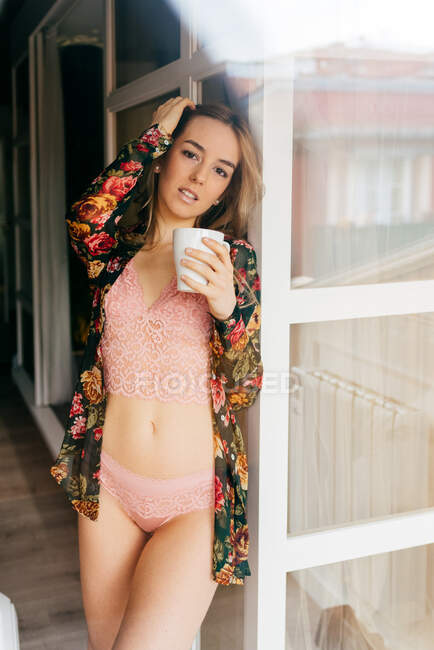 Attraente giovane femmina che tocca i capelli in lingerie elegante e accappatoio colorato mentre guarda la fotocamera con tazza di caffè dalle finestre a casa al mattino — Foto stock