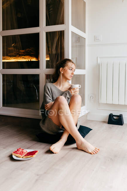 Sad young female in casual outfit sitting on floor with cup of coffee in hand and plate with toasts placed nearby and looking away pensively while spending morning at home — Stock Photo
