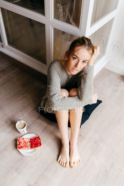 Зверху спокійна молода жінка в повсякденному одязі, дивлячись на камеру задумливо, сидячи на підлозі з чашкою кави і тостів під час сніданку вдома — стокове фото
