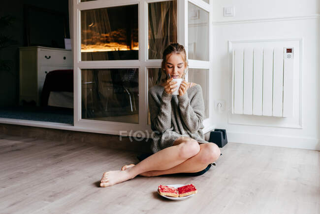 Mujer rubia joven feliz en traje casual sentado en el suelo con una taza de café en la mano y un plato con tostadas colocadas cerca mientras pasa la mañana en casa - foto de stock