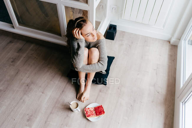 Dall'alto di calma giovane femmina in abbigliamento casual guardando la finestra pensieroso mentre seduto sul pavimento con una tazza di caffè e toast durante la colazione a casa — Foto stock
