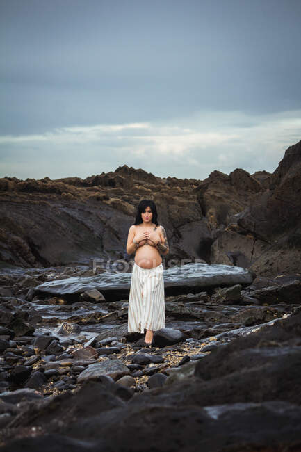 Sonhador topless morena bela mulher grávida em maxi saia de pé sobre rochas maravilhosas com fluxo e cobrindo o peito olhando para a câmera — Fotografia de Stock