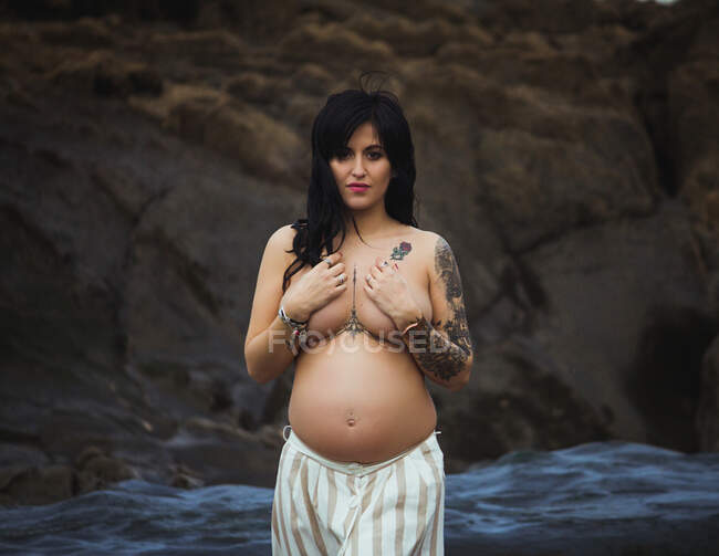 Rêve seins nus brune belle femme enceinte en maxi jupe debout sur des roches merveilleuses avec ruisseau et couvrant poitrine regardant caméra — Photo de stock
