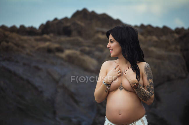 Мрійлива топлес брюнетка щаслива красива вагітна жінка в спідниці максі, що стоїть на чудових каменях з струмком і покриває груди, дивлячись геть — стокове фото