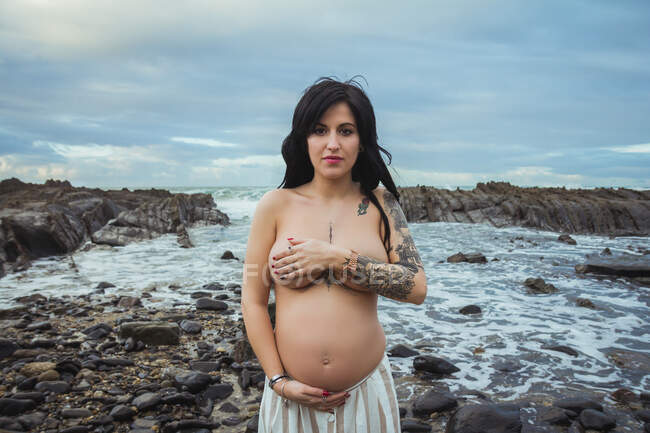 Sonhador topless morena bela mulher grávida em maxi saia de pé sobre rochas maravilhosas com fluxo e cobrindo o peito olhando para a câmera — Fotografia de Stock