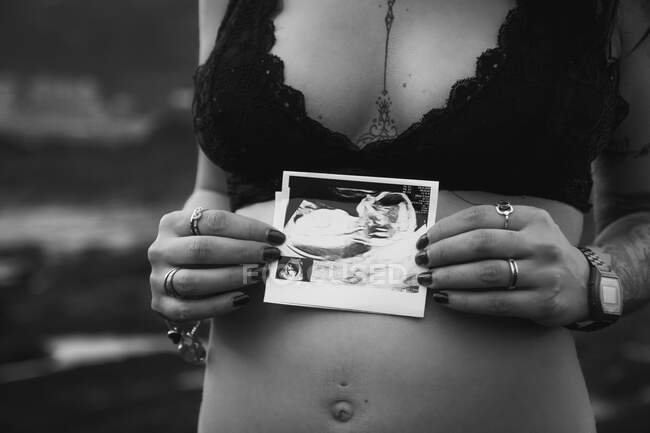 Tattooed stylish pregnant woman in bra holding sonogram picture on belly standing on majestic seacoast — Stock Photo
