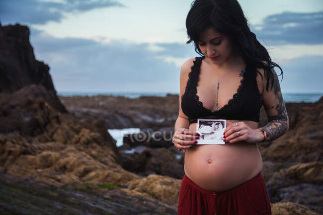 Татуйована стильна жінка під час вагітності тримає фотографію сонограми на животі, стоячи на величезному узбережжі з хмарним небом — стокове фото