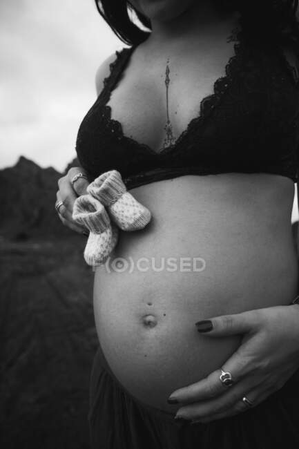 Black and white cropped faceless sensual pregnant woman touching belly and holding adorable baby shoes in nature — Stock Photo