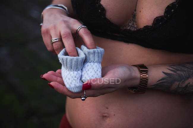 Cropped faceless pregnant woman touching belly and holding adorable baby shoes in nature — Stock Photo