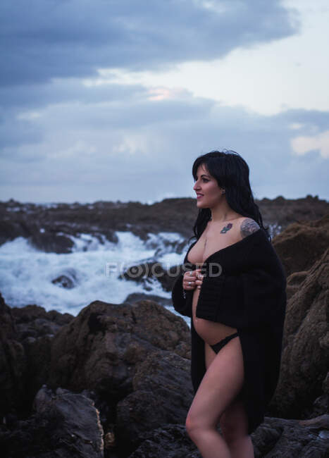Vista lateral de la mujer embarazada sensual feliz mirando hacia otro lado usando larga túnica de lencería ondulante caminando con el vientre desnudo en la costa rocosa en el día sombrío - foto de stock