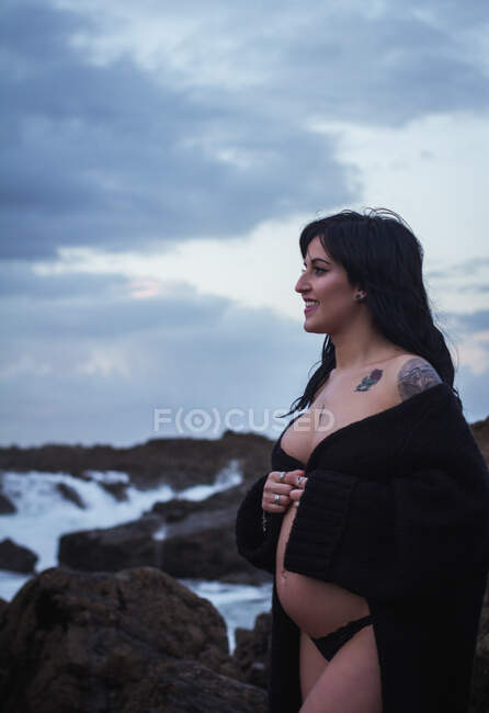 Vista lateral de la mujer embarazada sensual feliz mirando hacia otro lado usando larga túnica de lencería ondulante caminando con el vientre desnudo en la costa rocosa en el día sombrío - foto de stock