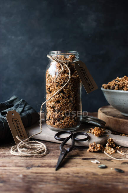 Glass jar of fresh millet and quinoa granola with label placed on wooden table near retro scissors — Stock Photo