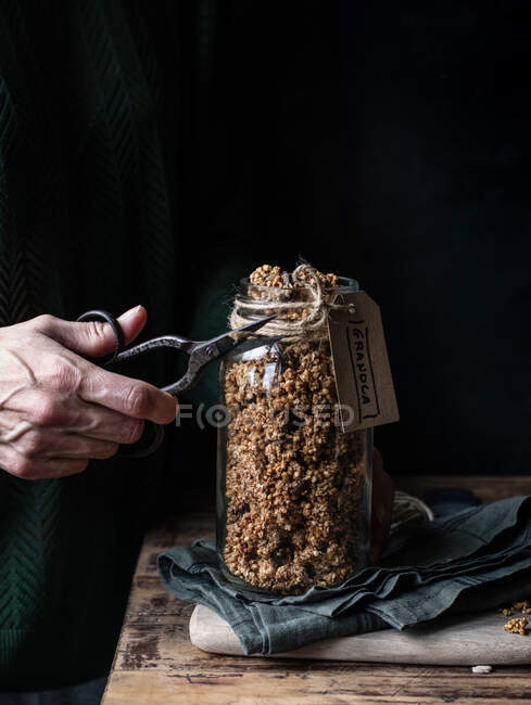 Unrecognizable person using retro scissor to cut rope with label from glass jar of millet and quinoa granola — Stock Photo