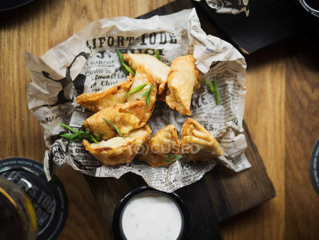 From above top view of delicious deep fried dumplings garnished with green herbs placed on paper served with white sauce on wooden table — Stock Photo