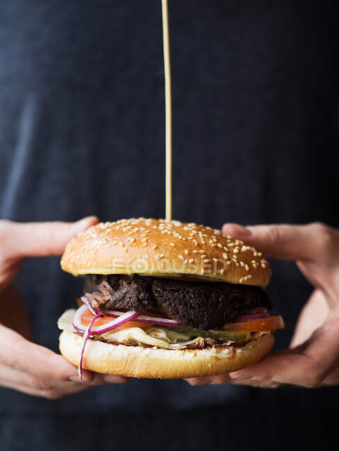 Crop pessoa anônima em desgaste preto segurando hambúrguer clássico com costeleta e legumes com queijo enquanto representa a indústria de fast food — Fotografia de Stock