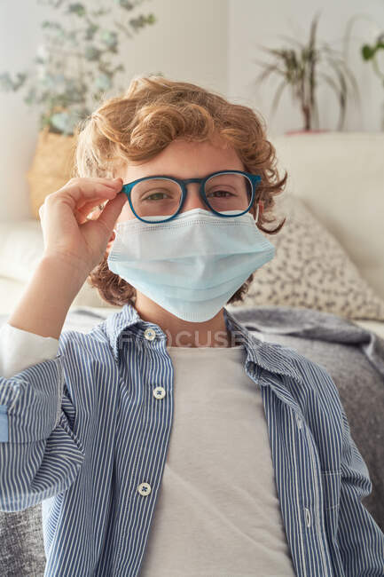 Умный мальчик в медицинской маске надевает очки, сидя за столом с ноутбуком и готовится к дистанционному обучению дома — стоковое фото