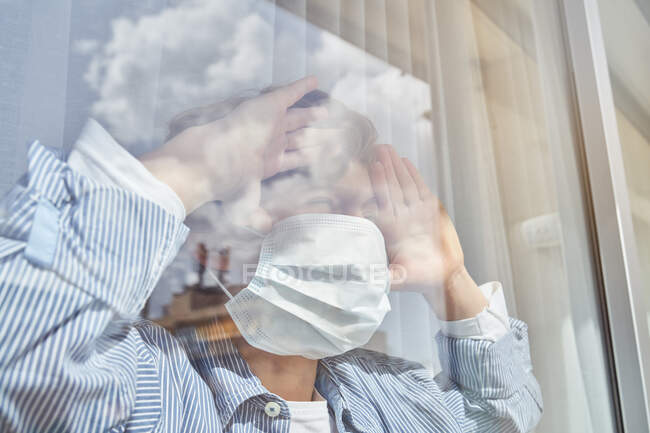 Мальчик в медицинской маске держит руку на стекле и смотрит в окно, оставаясь дома во время карантина — стоковое фото