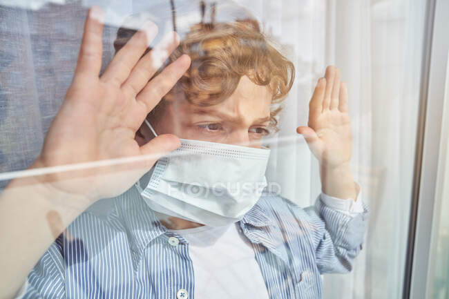 Хлопець у медичній масці тримає руку на склі і дивиться у вікно, перебуваючи вдома під час карантину. — стокове фото