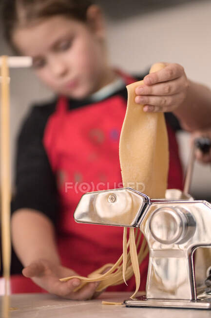 Маленька дівчинка в червоному фартусі використовує машину для макаронних виробів під час приготування саморобного спагеті на кухні — стокове фото