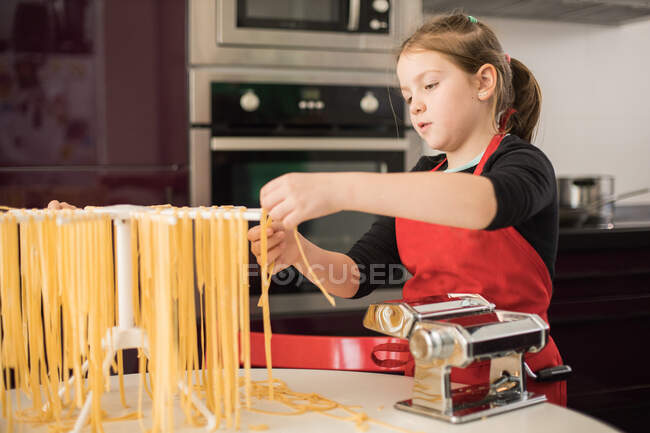 Grave menina pré-adolescente em avental vermelho pendurado macarrão cru no rack enquanto está à mesa com máquina de massas na cozinha — Fotografia de Stock
