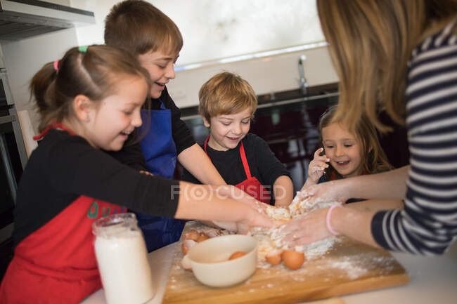 Crianças alegres em aventais de chef e mulher de colheita em pé em torno da mesa e amassar a massa enquanto prepara massas artesanais — Fotografia de Stock