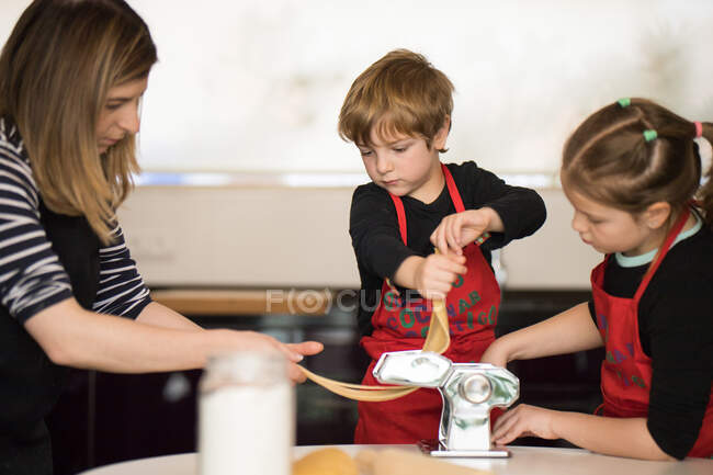 Menino e menina bonito em aventais de chef vermelho usando máquina de massas enquanto prepara macarrão com a ajuda de professora durante a aula de culinária — Fotografia de Stock
