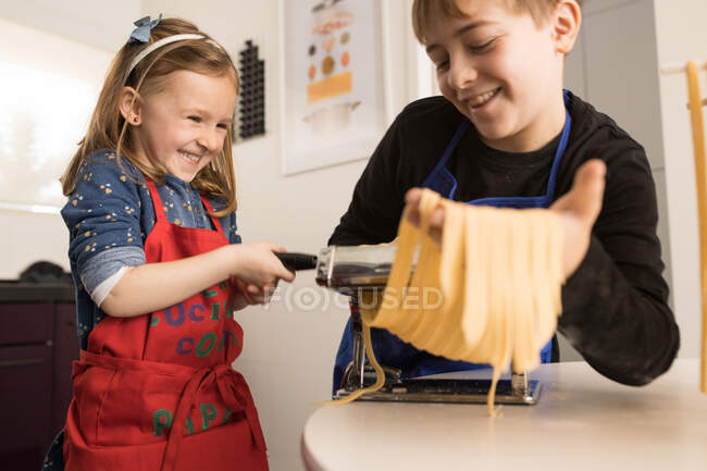 Une petite fille avec son frère aîné utilisant une machine à pâtes tout en préparant des nouilles maison dans la cuisine de la maison — Photo de stock