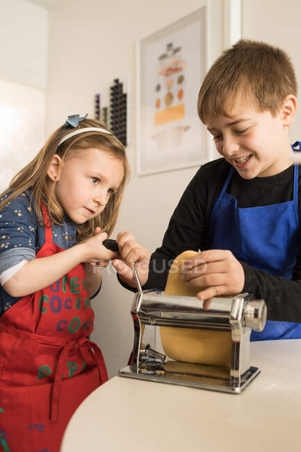 Маленькая девочка со старшим братом, использующая макароны во время приготовления домашней лапши на домашней кухне — стоковое фото