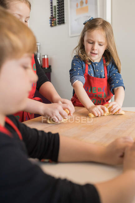 Cute little girl in chef apron kneading dough on wooden table while learning recipe for homemade noodles in cooking class with children — Stock Photo