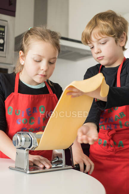 Una niña con su hermano usando una máquina de pasta mientras preparaba fideos caseros en la cocina casera - foto de stock