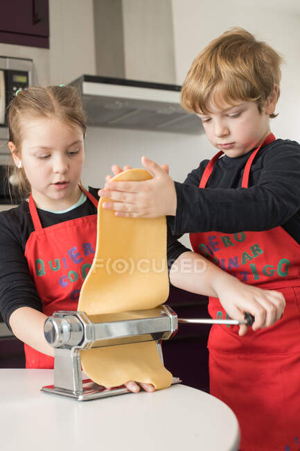Una bambina con suo fratello che usa la macchina per la pasta mentre prepara spaghetti fatti in casa in cucina — Foto stock