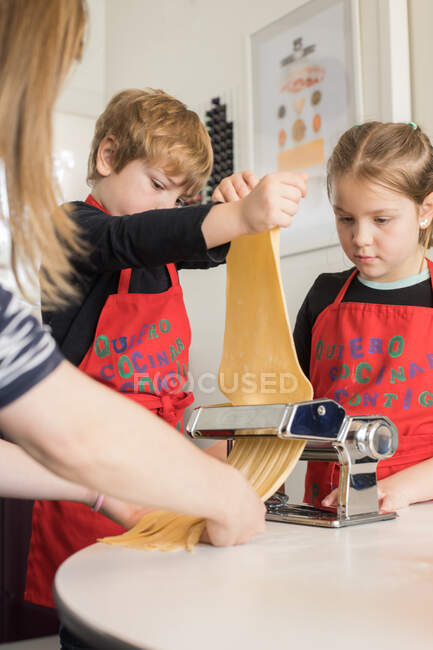 Una niña con su hermano usando una máquina de pasta mientras preparaba fideos caseros en la cocina casera - foto de stock