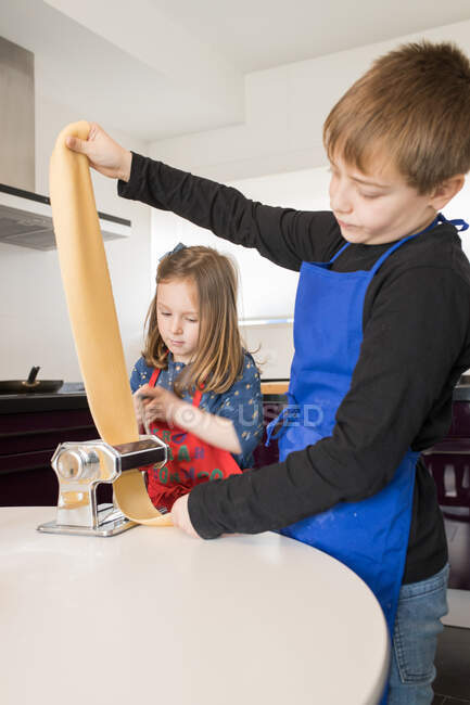 Маленька дівчинка з братом використовує макаронні машини, готуючи домашню локшину на домашній кухні — стокове фото