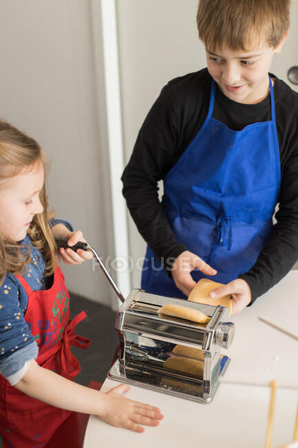Uma menina com seu irmão usando máquina de massas enquanto prepara macarrão caseiro na cozinha doméstica — Fotografia de Stock
