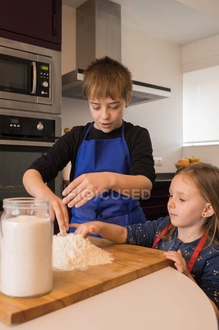 Cute little girl helping older boy in preparing dough while standing together at kitchen table with flour — Stock Photo
