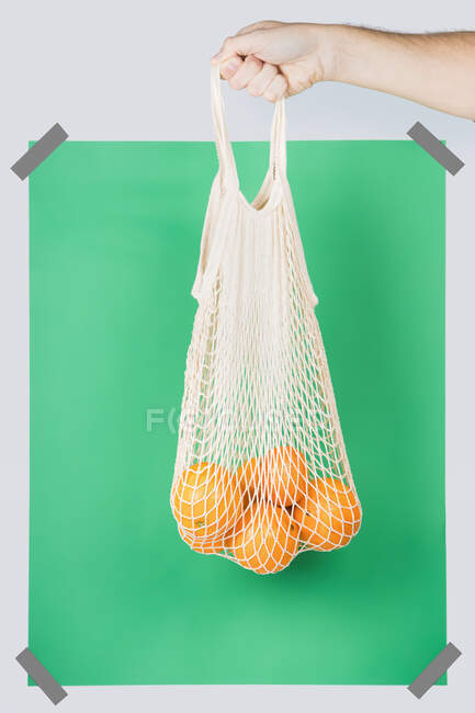 Unrecognizable person carrying net bag with ripe oranges against green rectangle during zero waste shopping — Stock Photo