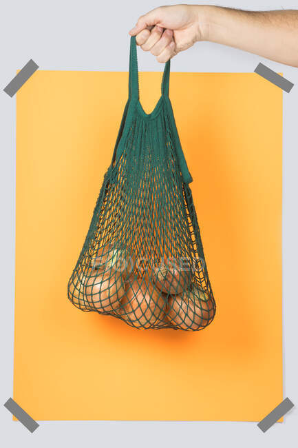 Persona anónima llevando bolsa de hilo verde con cebollas maduras contra rectángulo amarillo durante compras ecológicas - foto de stock