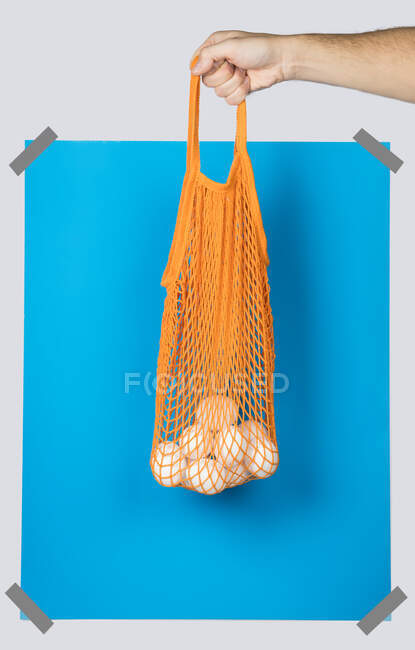 Personne méconnaissable portant un sac filet orange avec des œufs frais contre le rectangle bleu lors du shopping zéro déchet — Photo de stock