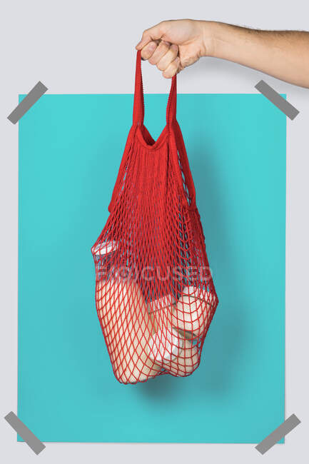 Anonymous person carrying red string sack with glass containers of dairy against turquoise rectangle during eco friendly shopping — Stock Photo