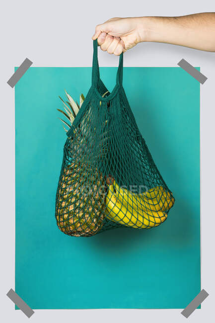 Sac filet pour personne méconnaissable avec ananas mûrs et bananes contre le rectangle bleu turquoise lors du shopping zéro déchet — Photo de stock
