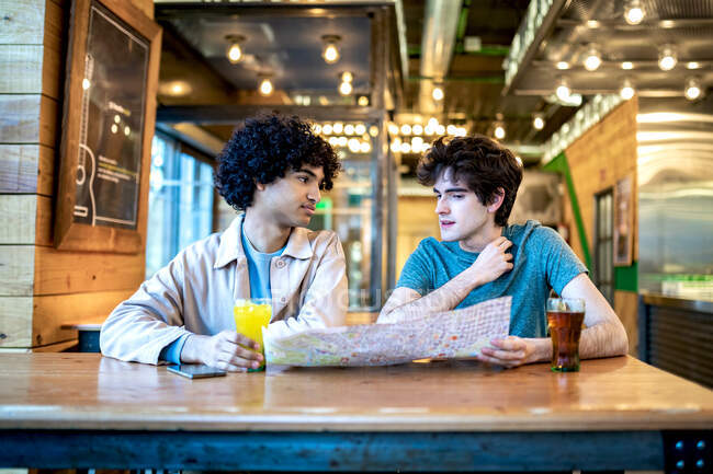 Multiethnische junge homosexuelle Männer mit Navigationskarte und frischen Getränken, die einander lächelnd beim romantischen Date am Cafétisch anschauen — Stockfoto