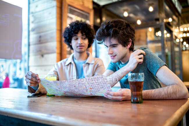 Мультиэтнические молодые гомосексуальные мужчины смотрят на навигационную карту направления и свежие напитки, улыбаясь, сидя за столом кафе во время романтического свидания — стоковое фото