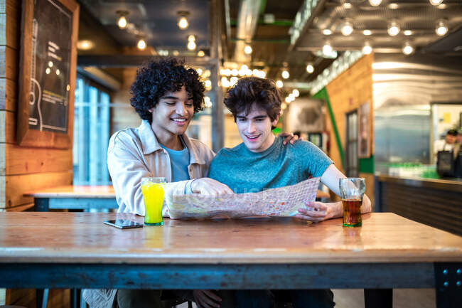 Багатоетнічні молоді гомосексуальні чоловіки дивляться на навігаційну карту напрямку і свіжі напої посміхаються, сидячи за столом кафе під час романтичного побачення — стокове фото