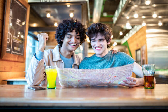 Multiétnicos jovens homossexuais olhando para direção mapa de navegação e bebidas frescas sorrindo enquanto sentado na mesa do café durante a data romântica — Fotografia de Stock