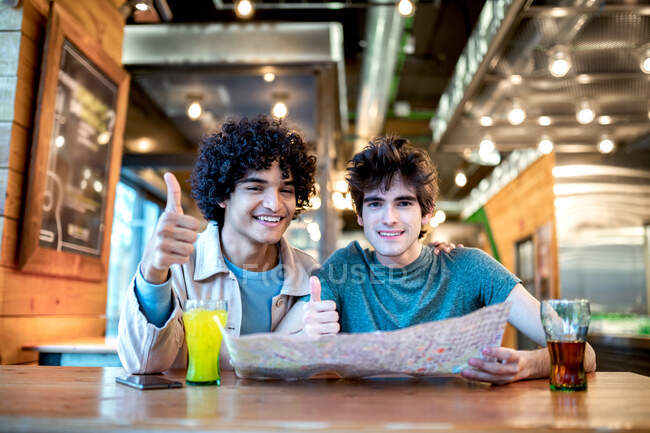 Багатоетнічні молоді гомосексуальні чоловіки з навігаційною картою напрямку та свіжими напоями посміхаються та показують жест великого пальця під час сидіння за столом кафе під час романтичного побачення — стокове фото