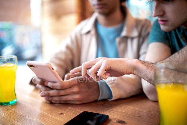 Cropped unrecognizable multiethnic young homosexual men browsing social media on smartphone and having fresh drinks smiling while sitting at cafe table during romantic date — Stock Photo