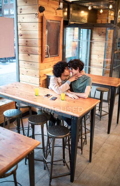 Зверху схвильовані етнічні чоловіки обіймаються і цілуються один з одним на столі і сміються під час романтичного побачення в сучасних кафетеріях — стокове фото