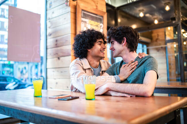 Vista lateral de hombres étnicos emocionados abrazándose con los ojos cerrados en la mesa y riendo durante la cita romántica en la cafetería moderna - foto de stock